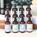Twelve bottles of high-end lotions by Whole Self Aromatherapy atop a piece of wood with spa towels on one side and a flower on the other.
